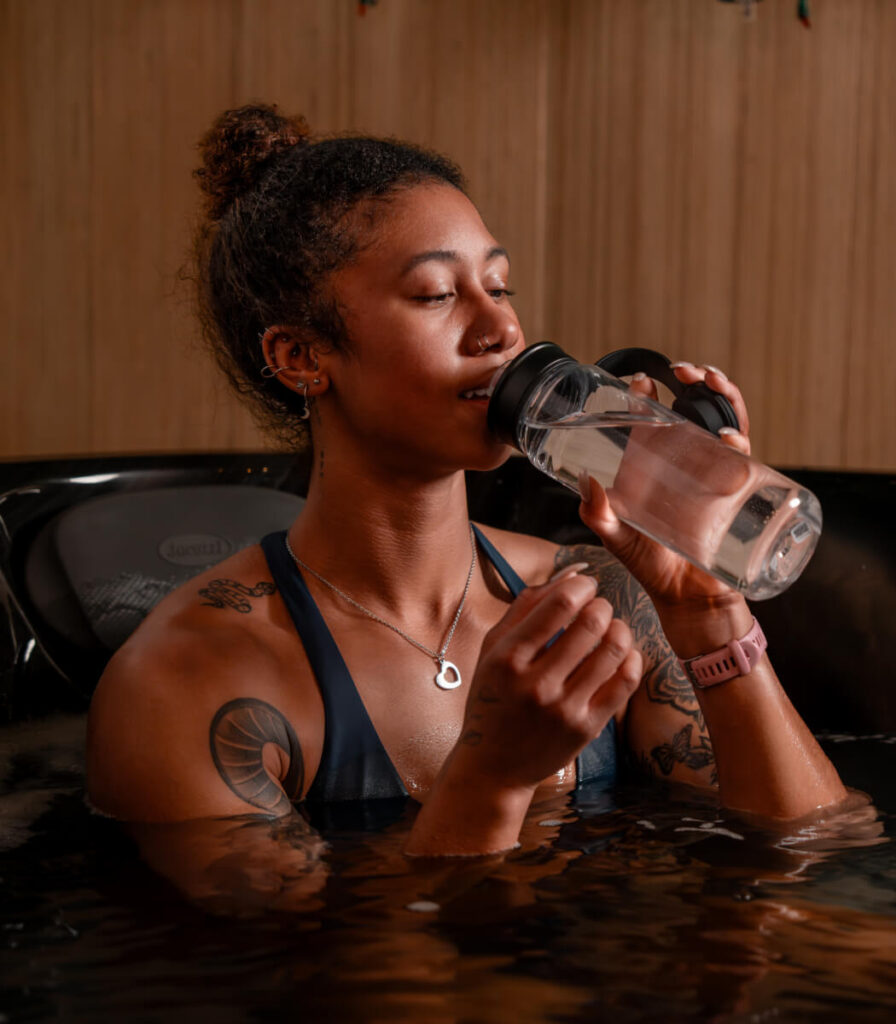 A woman sitting down while drinking water from a bottle and doing Warm Water Hydrotherapy.